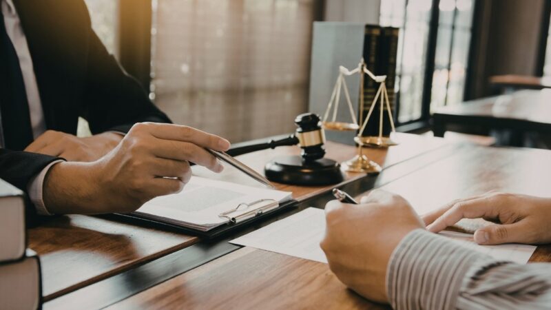 Advokat u kancelariji pokazuje dokument klijentu dok ovaj potpisuje ugovor za stolom